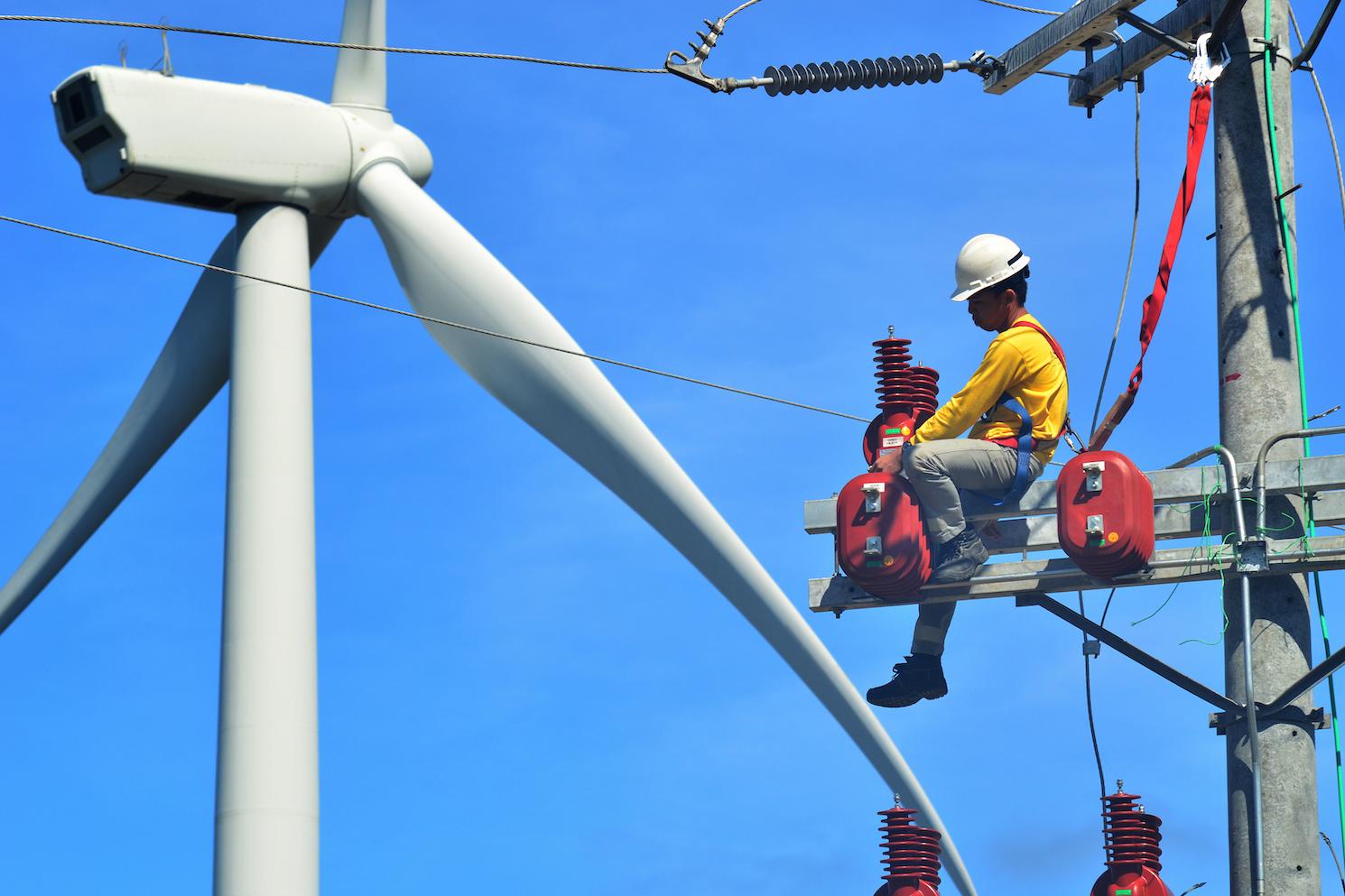 A technician sits on his high post repairing an electrical pole in front of a wind turbine.