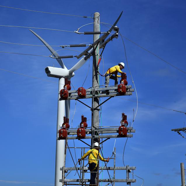 Technicians repairing an electrical pole in front of a wind turbine.