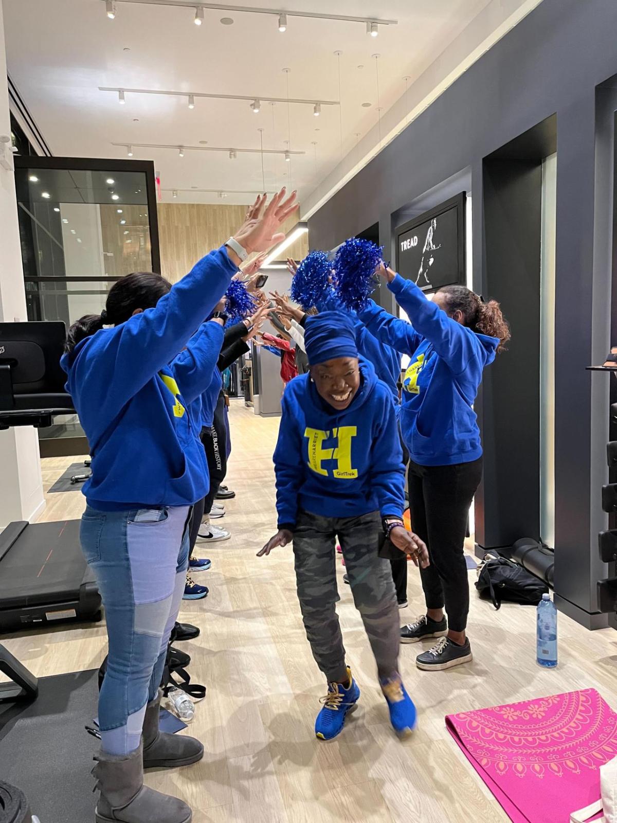 Women in blue sweatshirts holding hands up to create a tunnel for another woman in a blue sweatshirt with a yellow "H" on it to walk through