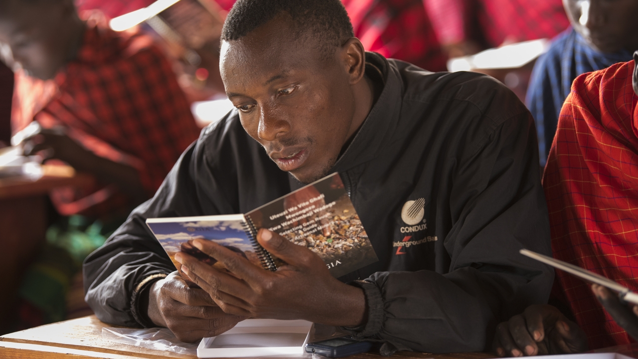 This ruby miner from Longido, Tanzania, is captivated by the lessons in recognizing quality and value in rough gem material. Selecting Gem Rough: A Guide for Artisanal Miners, authored and produced by GIA, is distributed for free to miners in East Africa. The booklet comes with a translucent white tray where miners can sort and examine rough. Photo by Robert Weldon/GIA (c) 2019