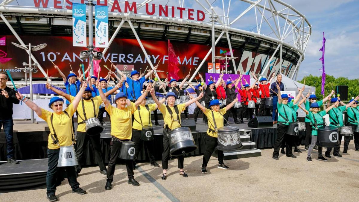 Groups of people in matching colored shirts playing instruments in a stage outside a stadium "Westham United"