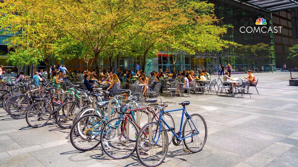 A paved courtyard with a row of bicycles. People eating and working at small tables under the shade of a large tree. A building with "Comcast" sign behind them.