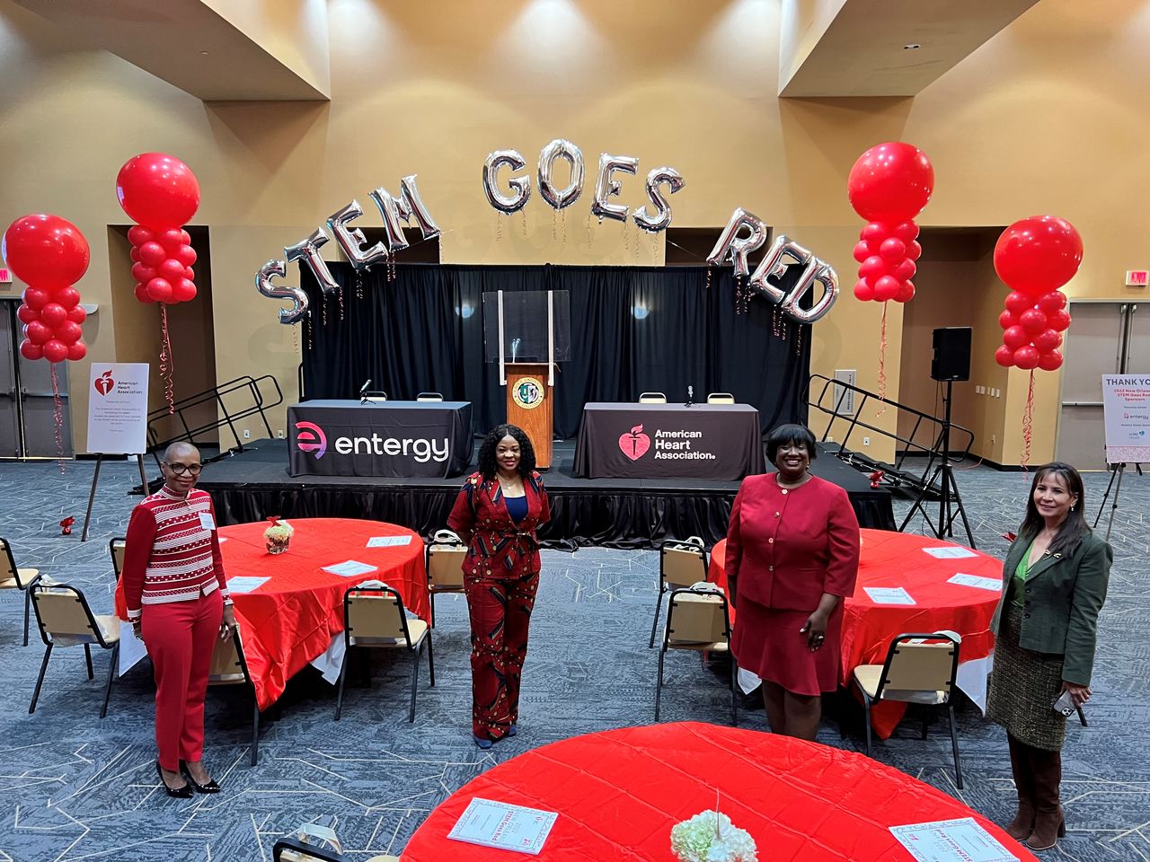 Four women standing in front of a stage featuring balloons that read "STEM GOES RED"