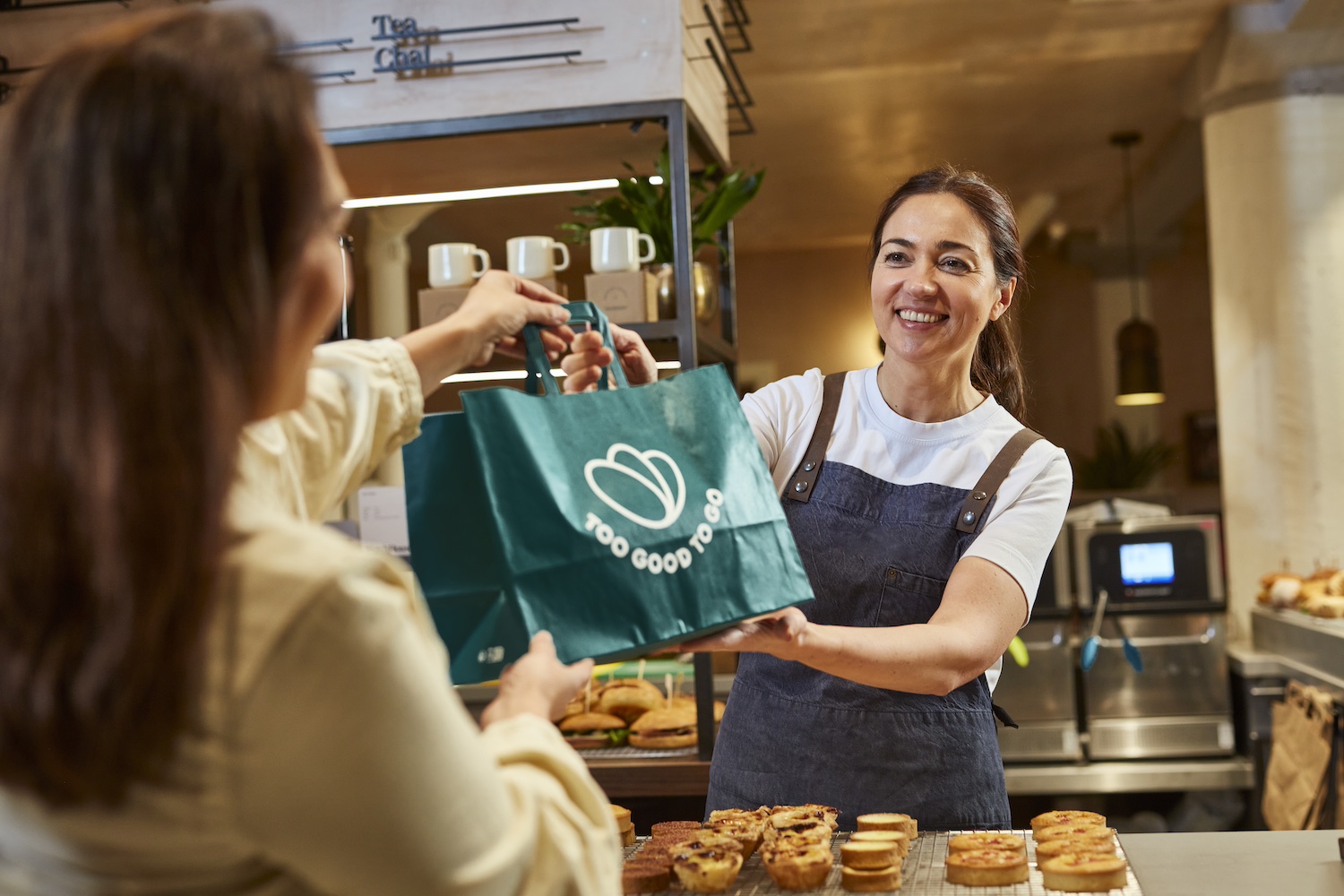 A person picks up a Too Good to Go surprise bag of food at a bakery. 
