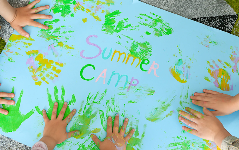 Children paint handprints on a sign with the inscription "Summer Camp"