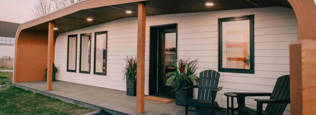 Exterior of the 3D printed home's front porch. Two chairs and a table to the side, plants on the sides of the doorway.