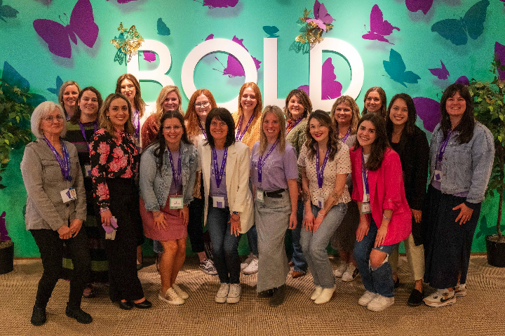 18 Bath & Body Works associates stand smiling in front of a seafoam green and purple wall decorated with purple butterflies and the word “bold” in white capital letters.