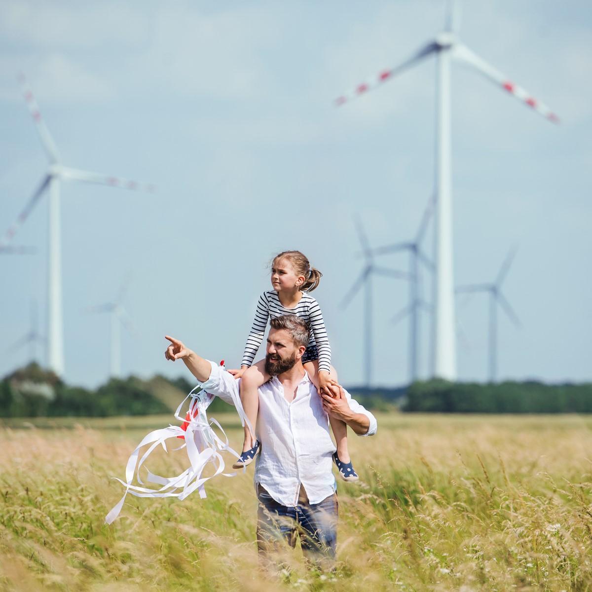 An adult with a child on their shoulders pointing into the distance. Wind turbines in a field behind them. 