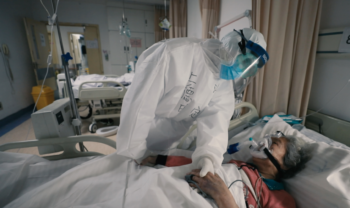 person in full PPE holding hands with a patient in bed