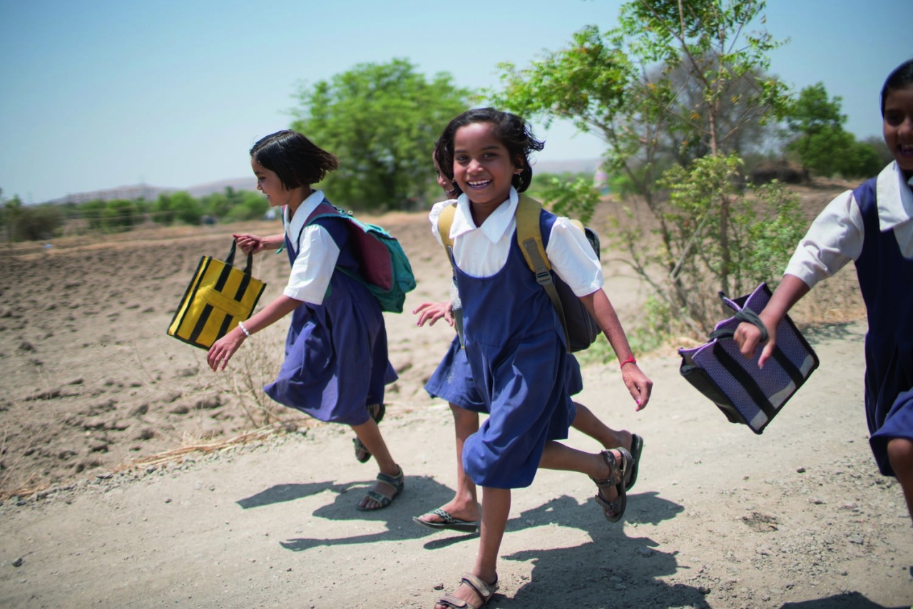 Four smiling children carrying backpacks running along a dirt road