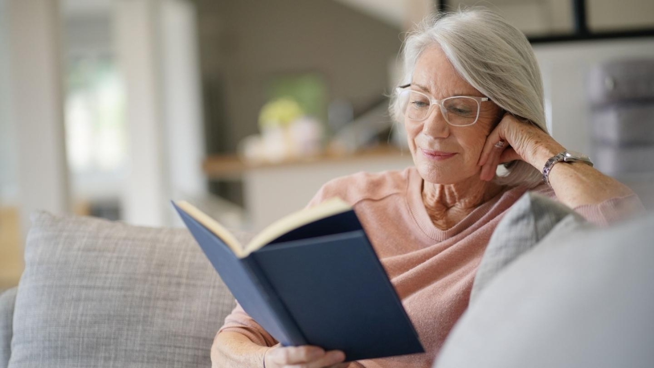 senior in a pink shirt on a gray couch reading from a blue book