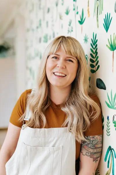 Ally Watson, smiling, leaning on a wall with painted plants.