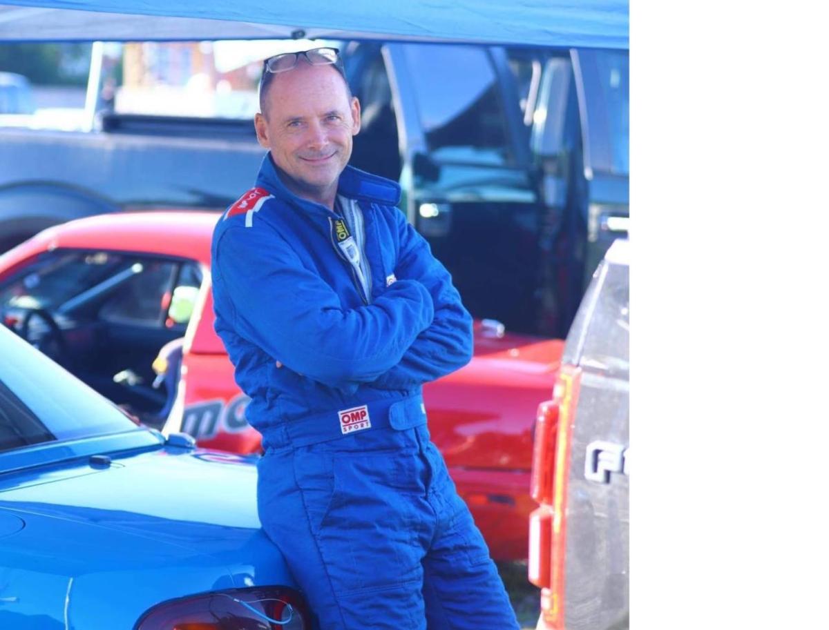 Andy O'Brien in a race suit, leaning on a car.