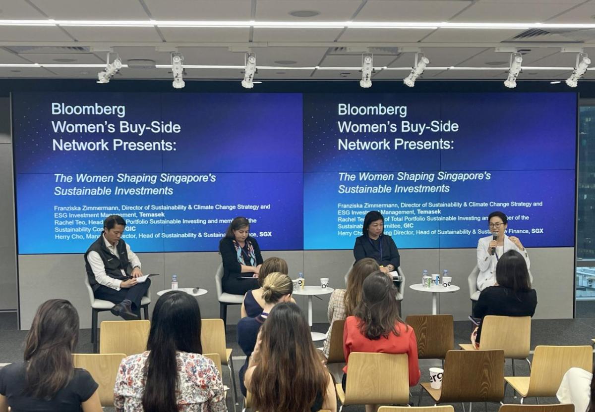 Four people on a stage, a small white table next to each of them. A small audience in front of them. Two digital projection screens behind them displaying "Bloomberg Women's Buy-Side Network Presents: The Women Shpaing Singapore's Sustainable Investments."