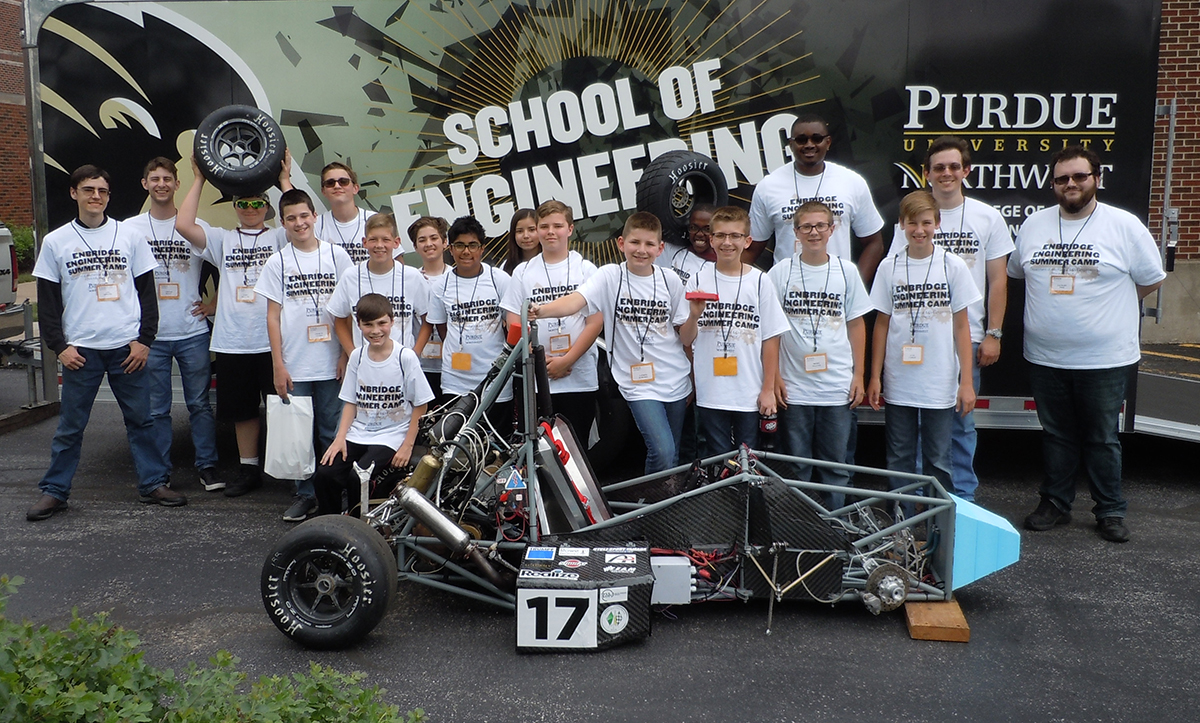 Large group of students and adults stand behind a go-cart type vehicle. All wearing the same 'school of engineering' t shirt
