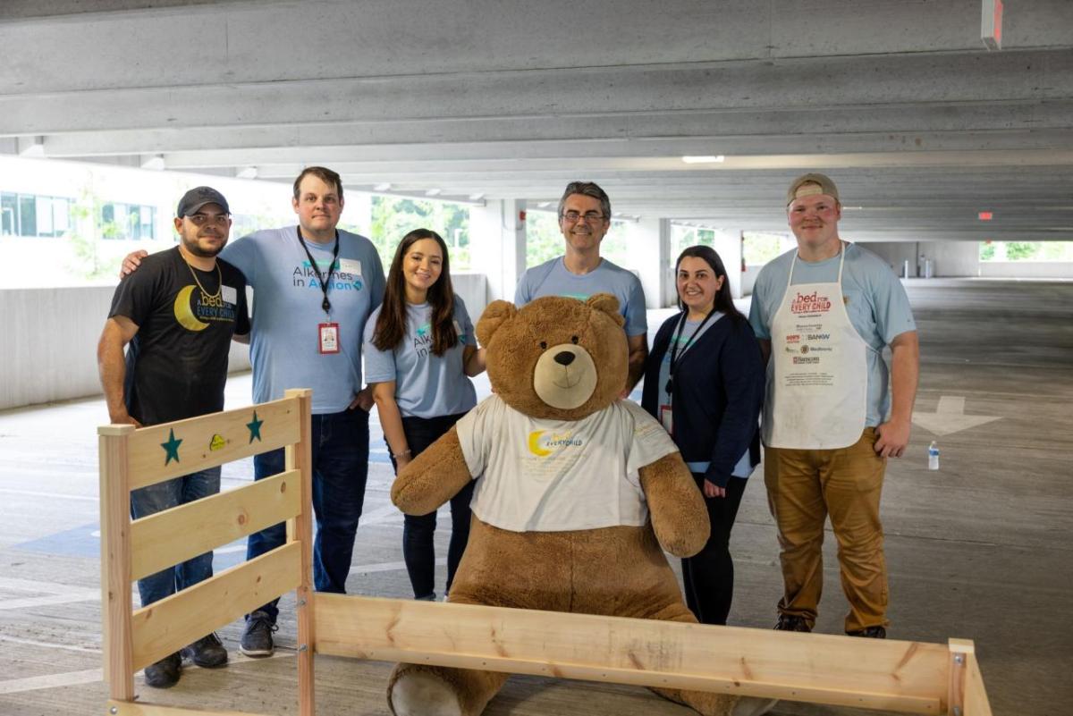 A group of volunteers posed with a large teddy bear and a bed frame.