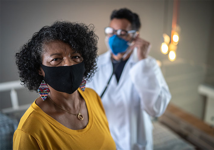 A medical professional using a stethoscope on a patient who is looking at the camera. Both are wearing protective masks.