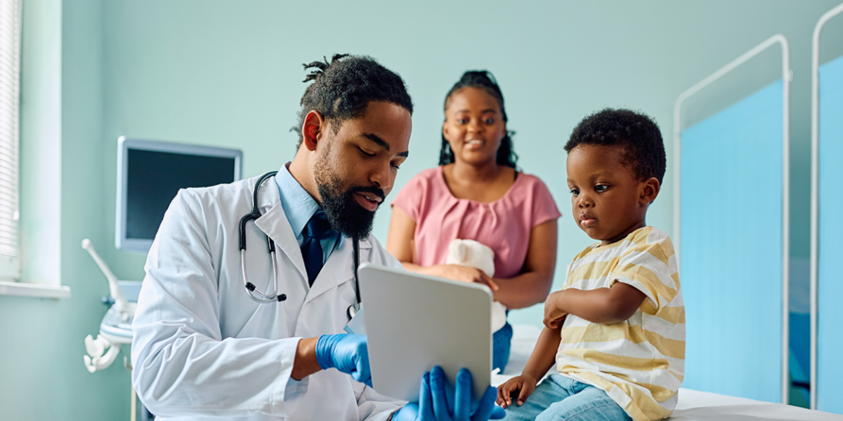 A doctor points to a tablet as a small child looks on. An adult smiling in the background