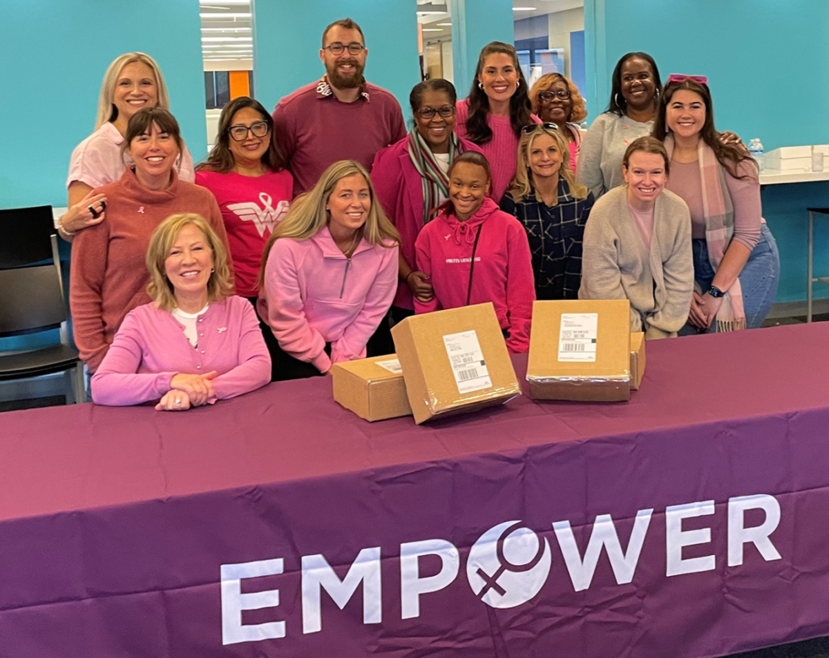 large group of volunteers dressed in pink pose behind a table that has an EMPOWER employee resource group tablecloth on it. On the table are Hope Kits.