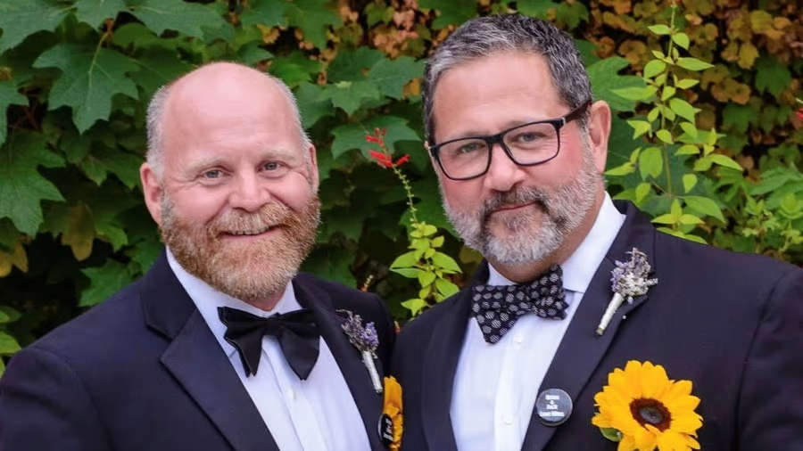 Brian Johnson and husband in suits, bowties and sunflowers in their front pockets.