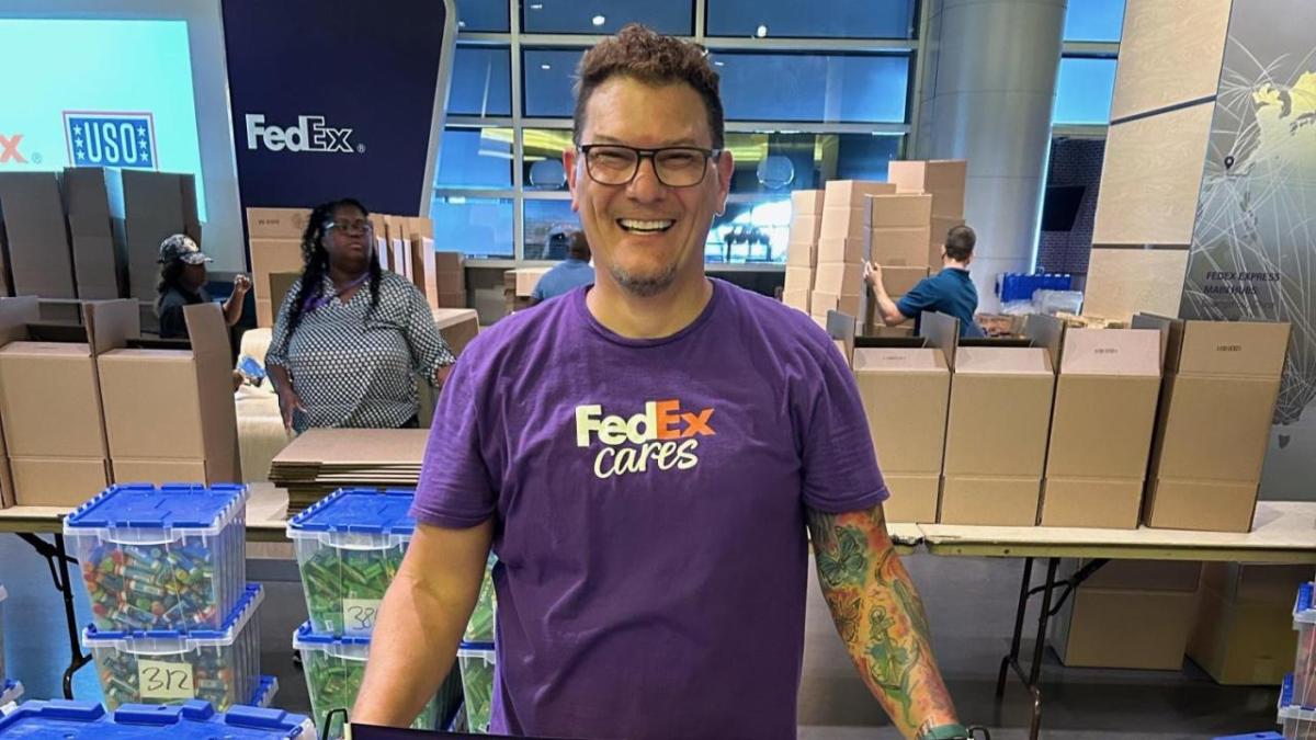 Brock Carlson, Manager- Talent Acquisition FedEx Logistics, stood in front of boxes wearing a purple fedEx cares photo