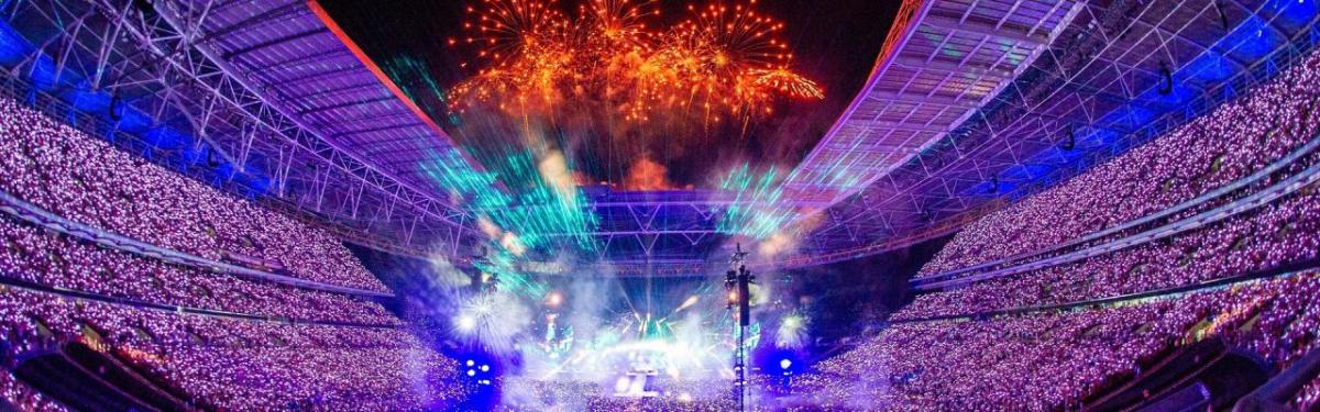 A huge stadium filled with spectators, a stage central is brightly lit, and orange fireworks fill the sky.