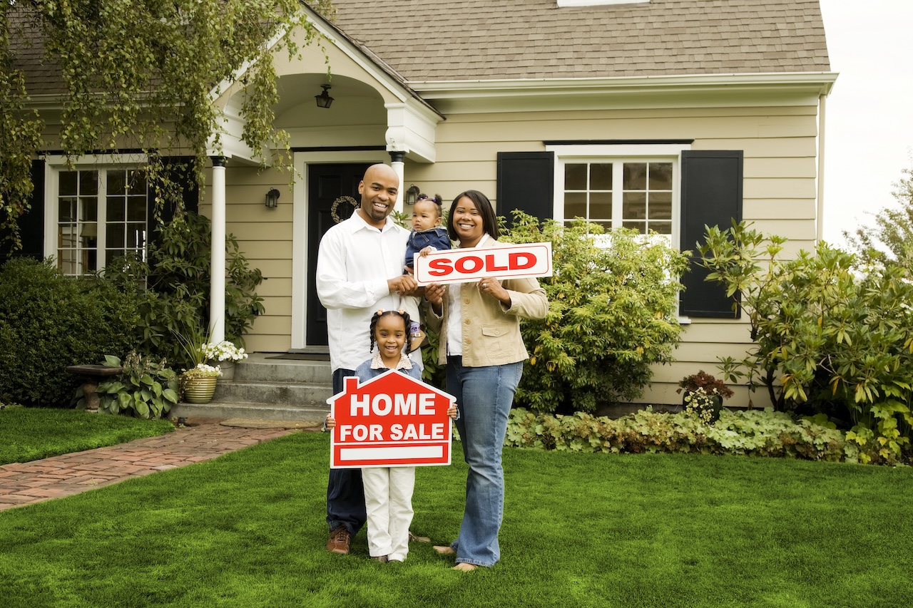 Young family of four in front of their new home holding a "sold" sign.