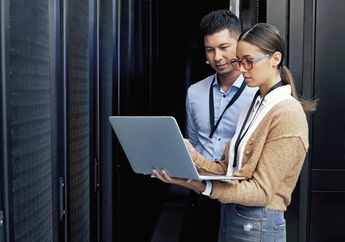 Two people in a server room and viewing a laptop.