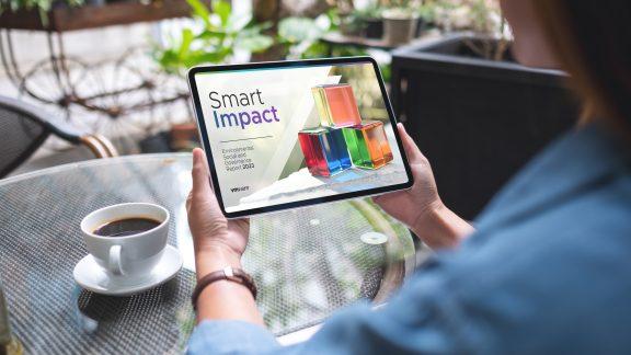 A person holding a tablet device with "Smart Impact" on the screen. A cup of coffee on the table in front of them.