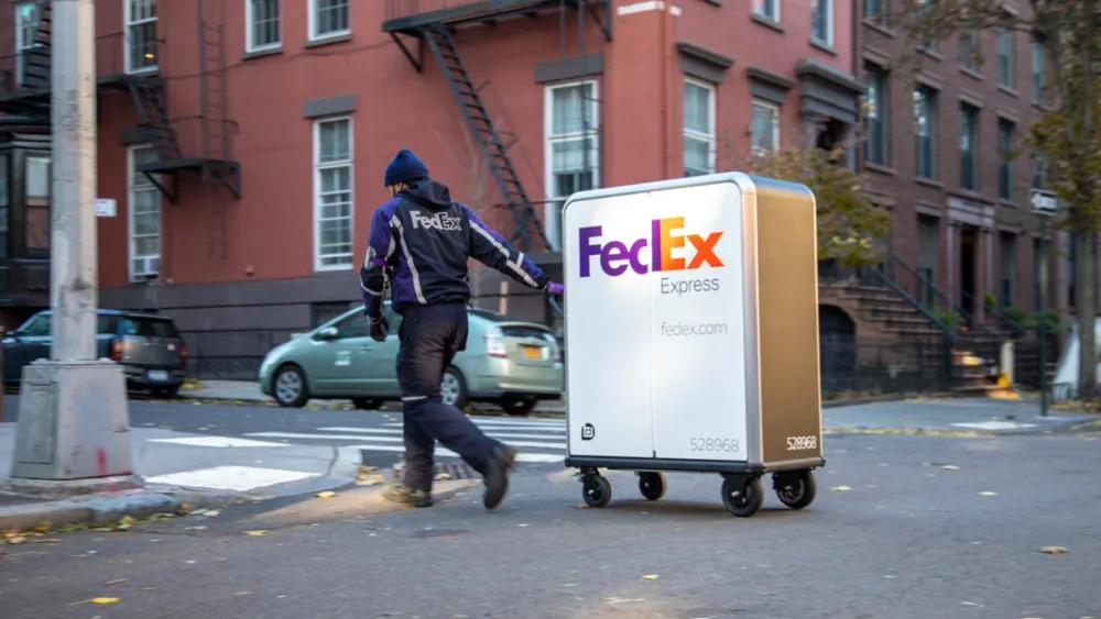 A person in FedEx uniform pulling a tall cart with FedEx Express logo on it crossing a street.