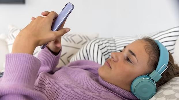 Teenage girl lying down wearing headphones and reading her phone.