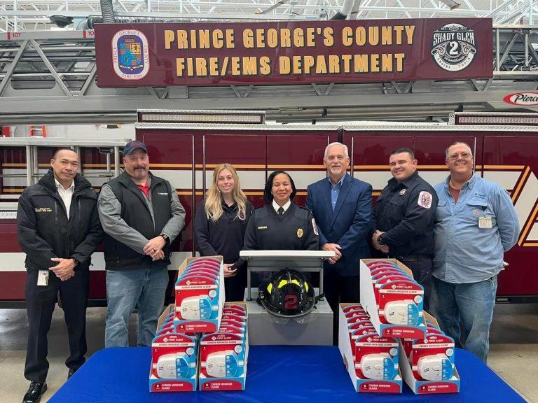 A line of people standing in front of a fire truck with a banner "Prince George's county fire/ems department". A table with smoke detectors in front of them.