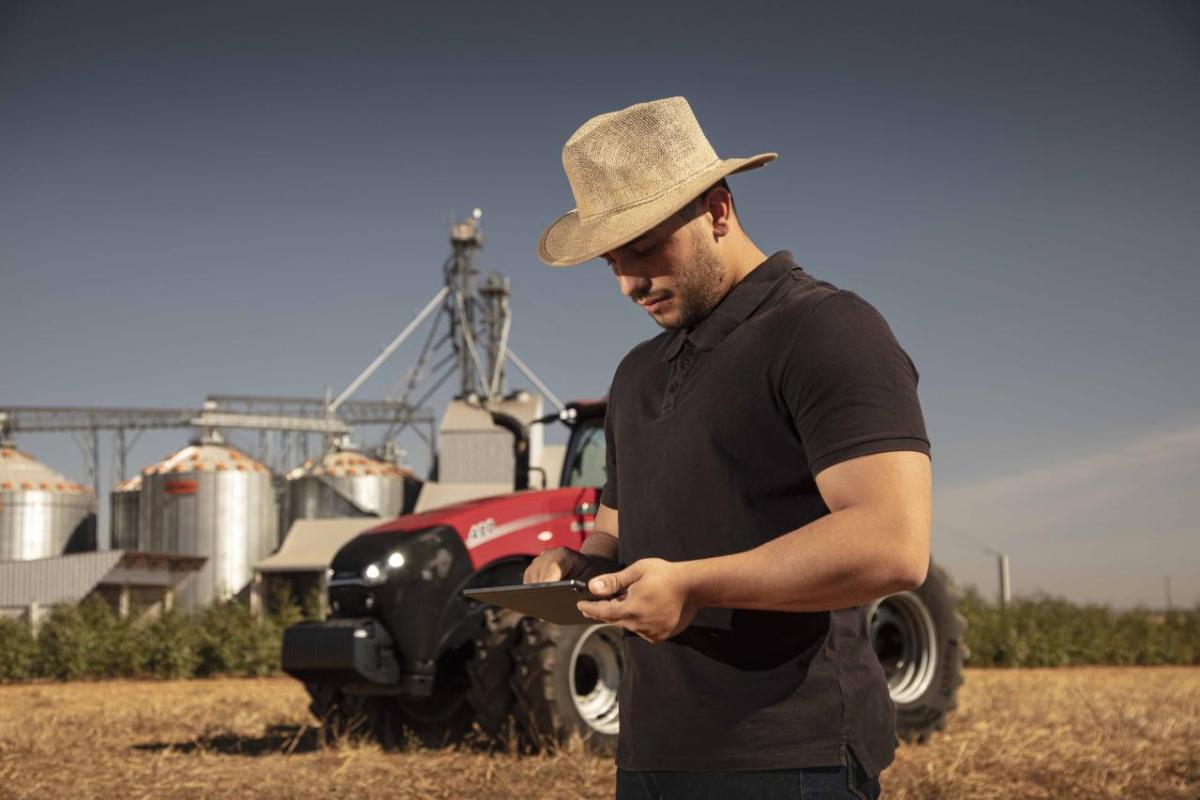 A farmer in a field looking at a tablet device. A red tractor and storage tanks behind them.