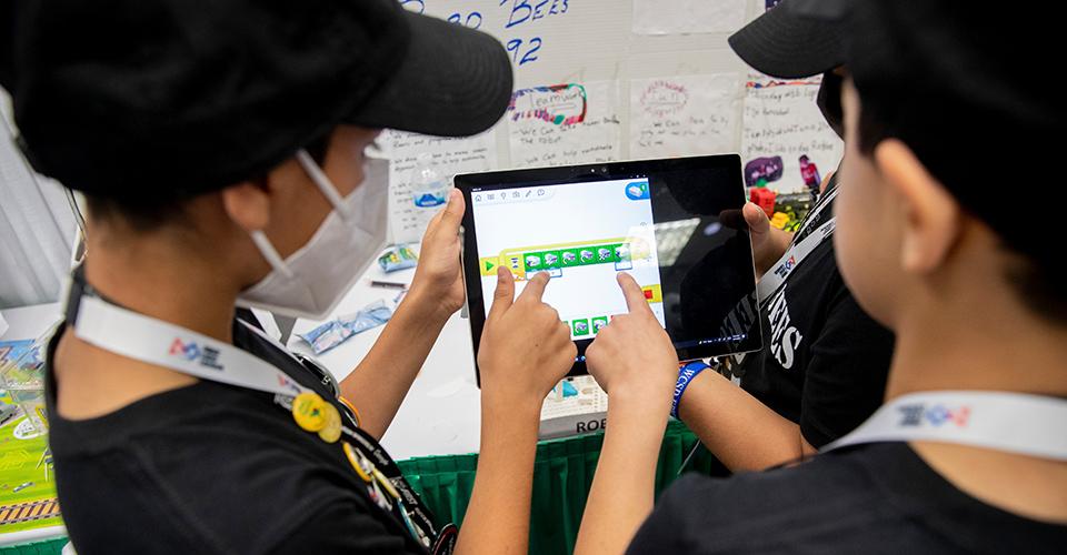 Children pointing at the same tablet, wearing matching lanyards and hats.
