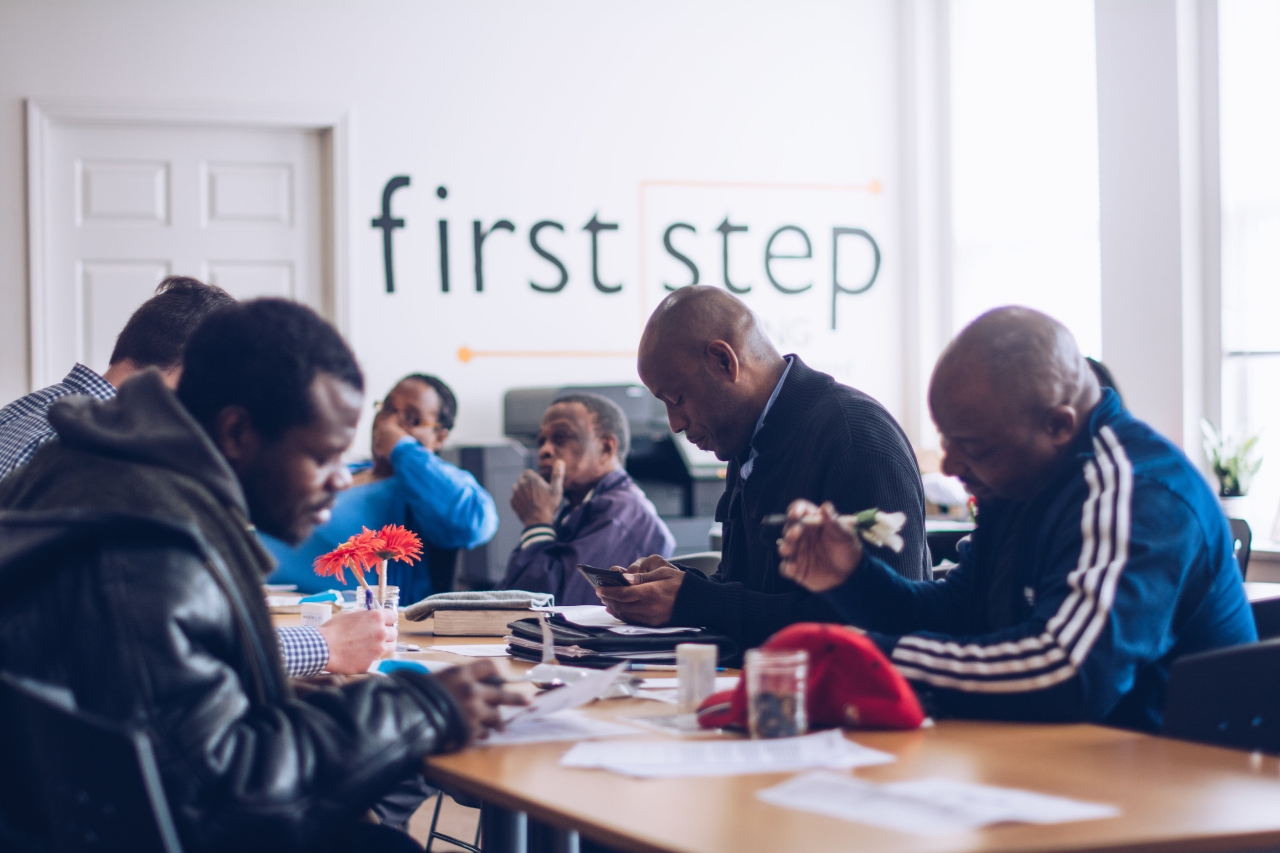 a group of people sitting at a table, filling out forms. "first step" banner in the background