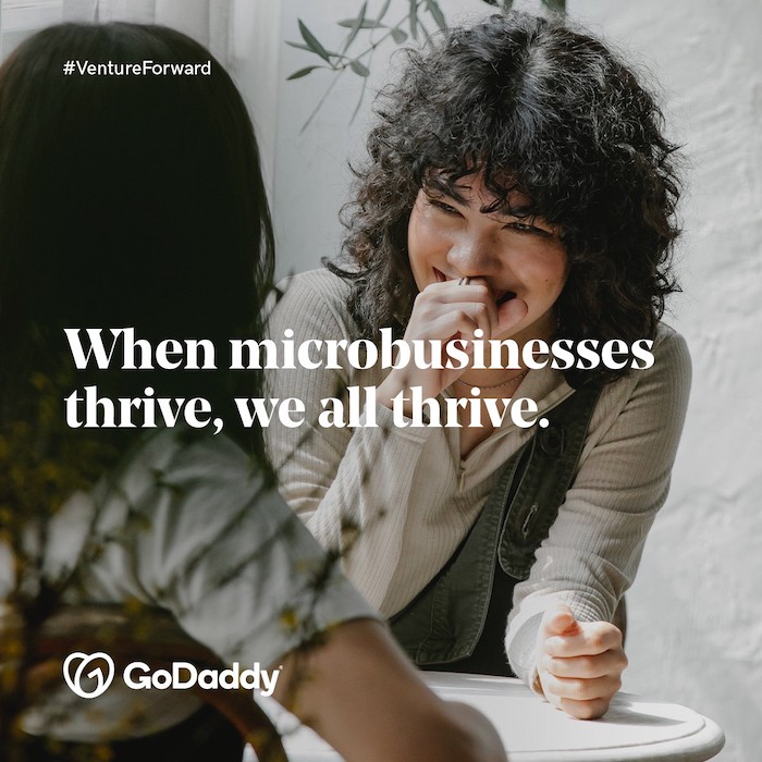 GoDaddy: Venture Forward. When microbusinesses thrive, we all thrive. Photo of two women seated at a small table.