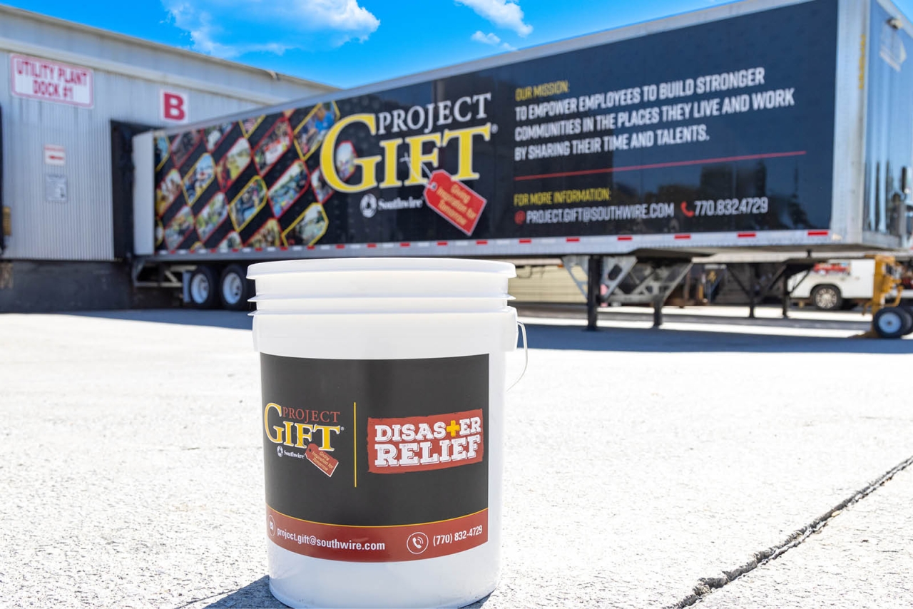 a white bucket with "GIFT disaster relief" label. A trailer in the background with "Project GIFT" on the side