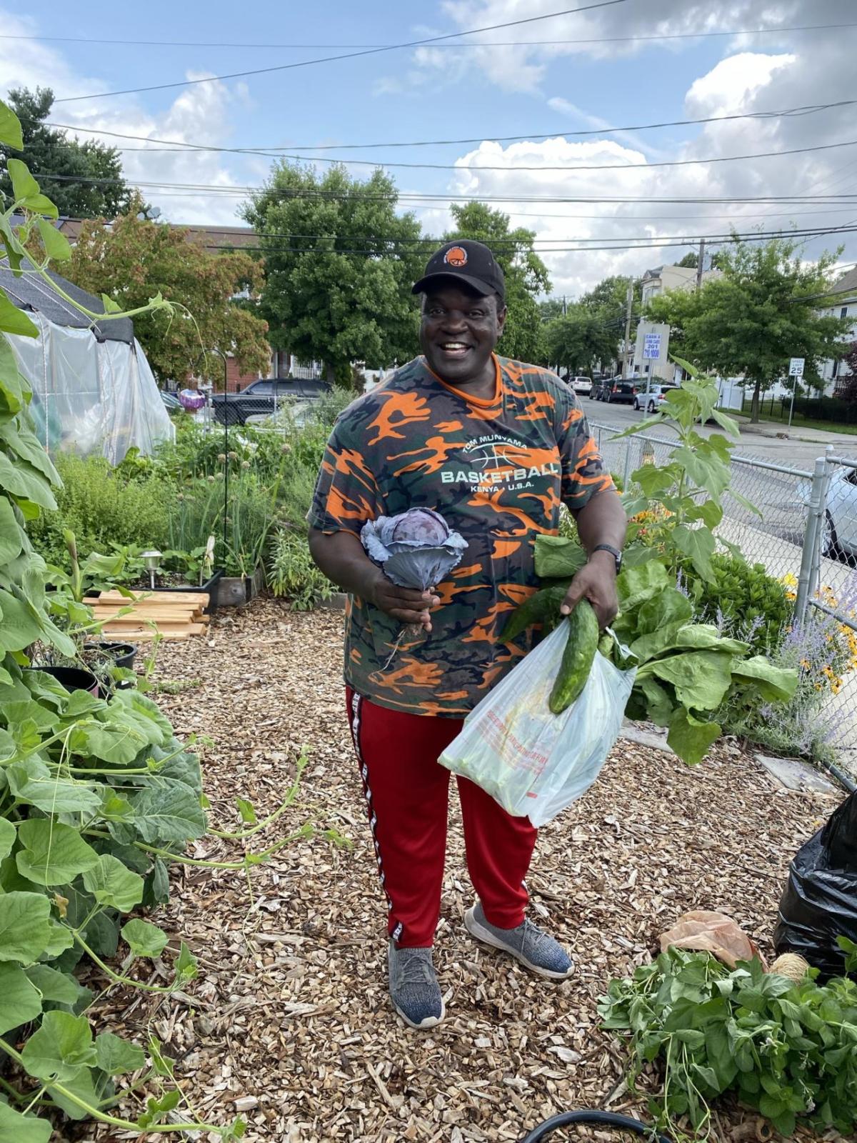 A person holding fresh produce, smiling. They're standing on a mulch path in a garden, a car goes by on the street next to them.