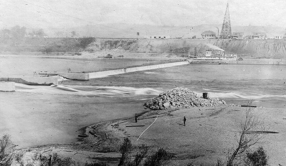 old aerial photo of a river with a dam across it. a building on the distant hill.