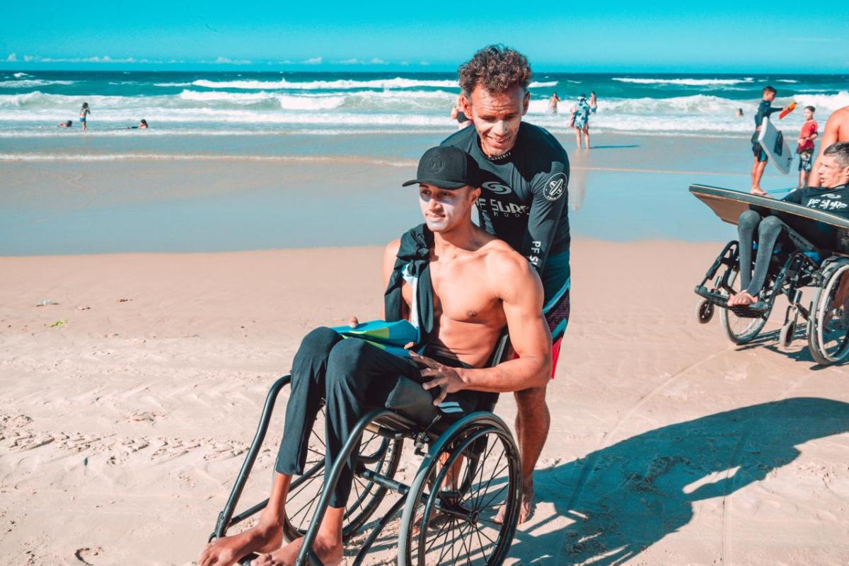 A person in a wet suit helping another in a wheelchair on a beach.