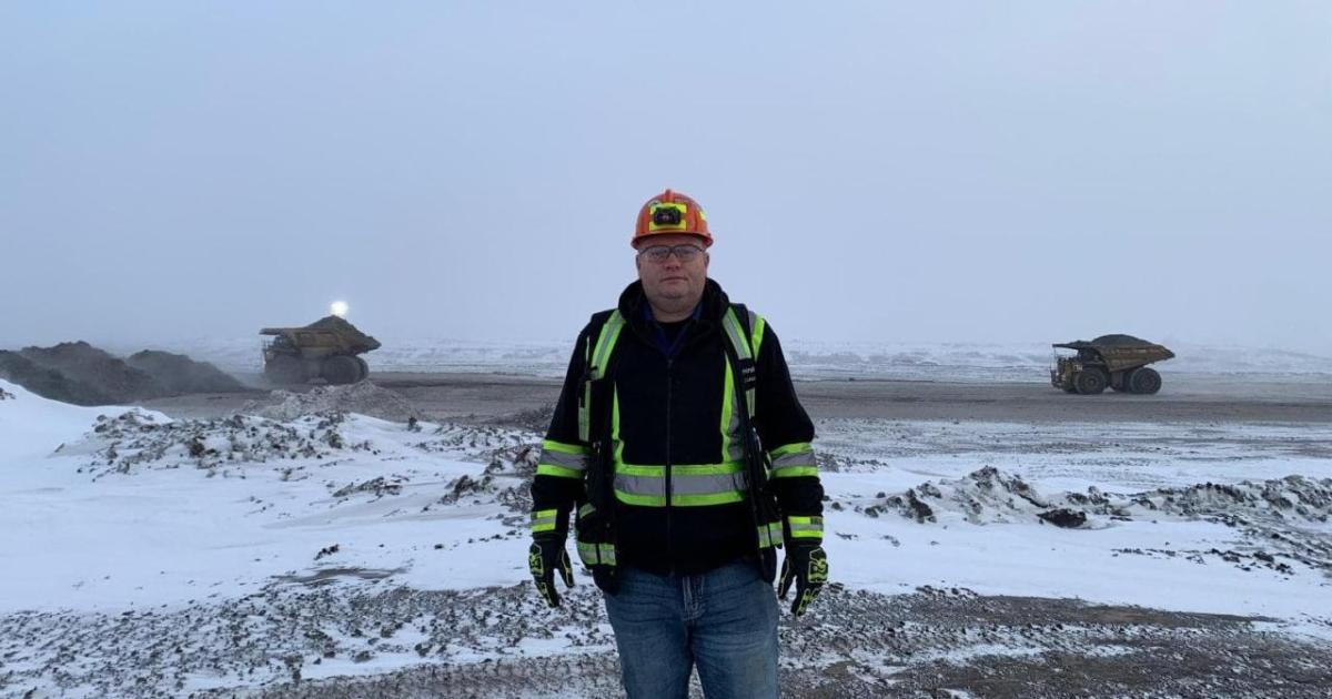 Jason Mercer in protective and high-vis jacket and hard hat outside in a snow covered quarry, dense fog behind them and two large mining dump trucks.