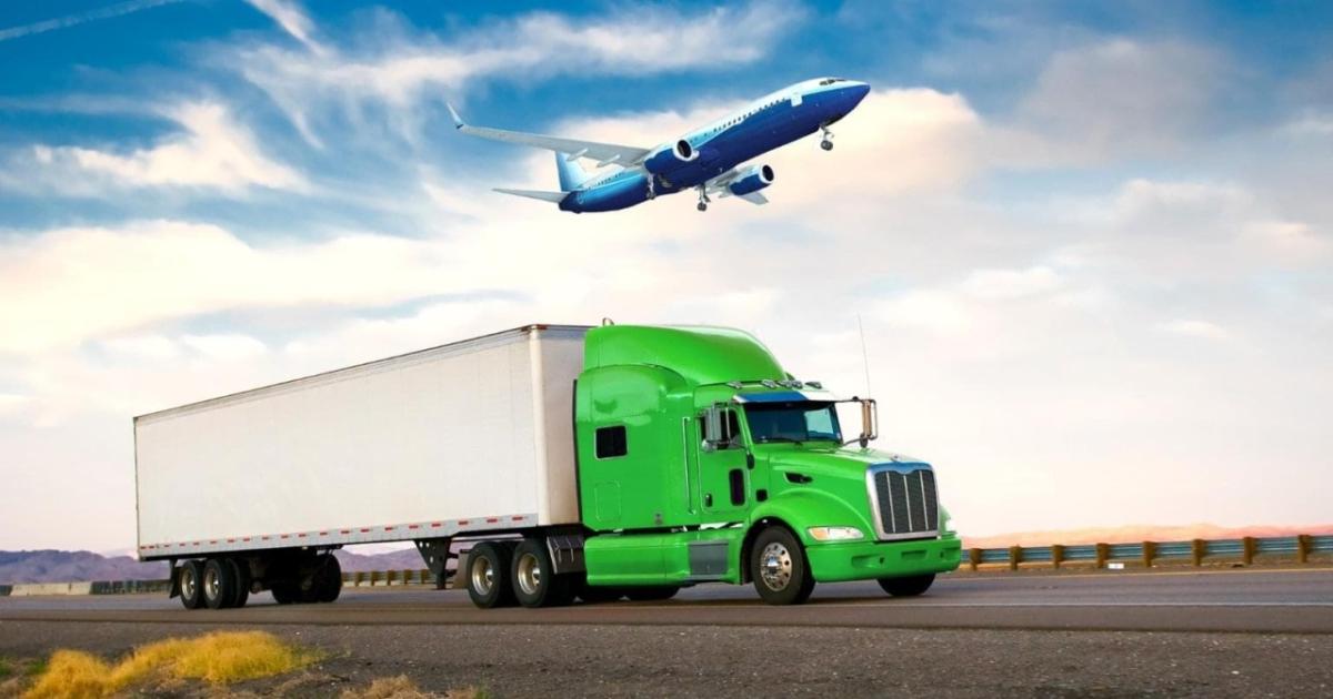 A green semi truck on the road and a jet plane in the sky above it.