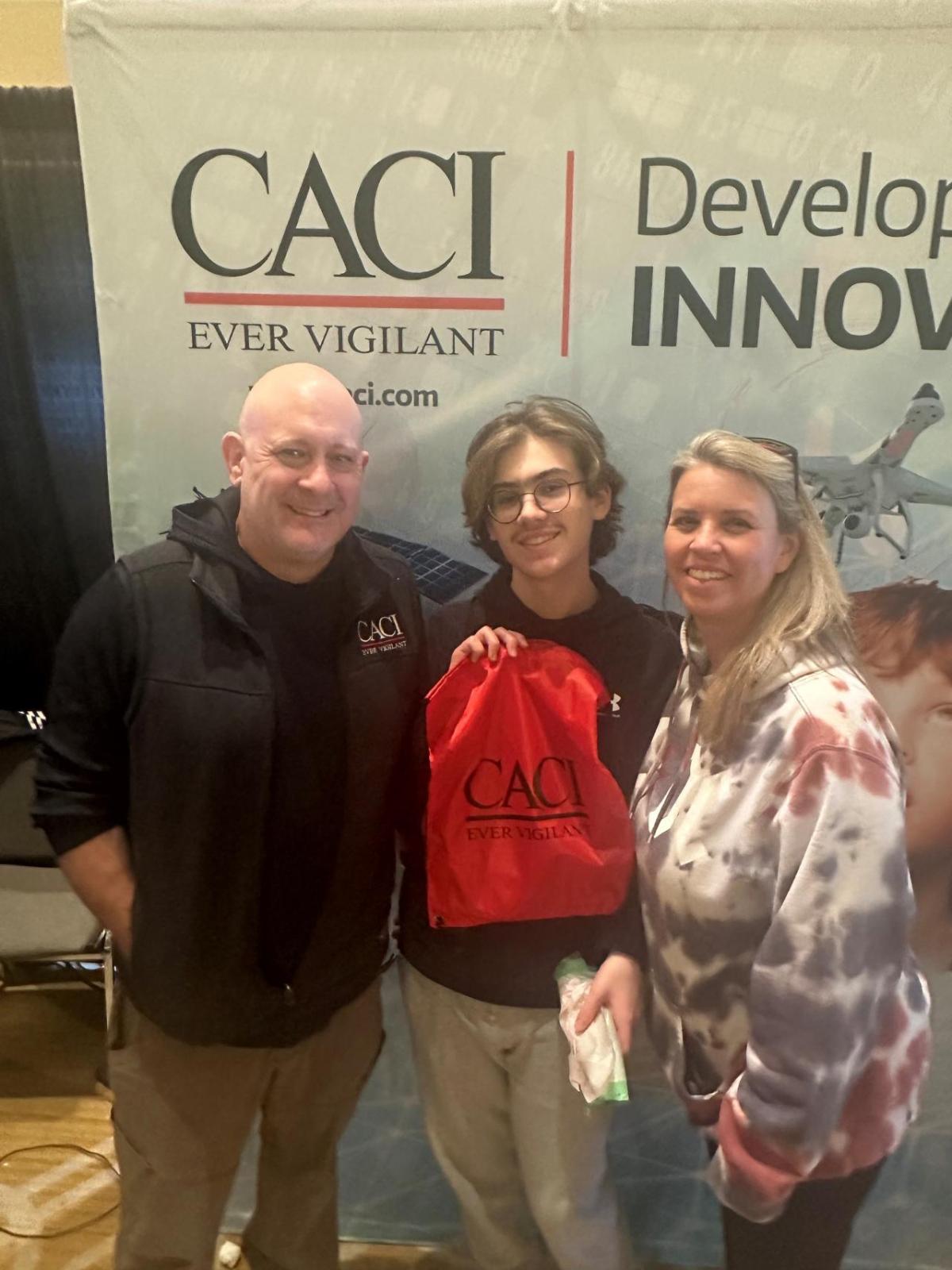 A group of three pose in front of a CACI banner, holding a CACI bag.