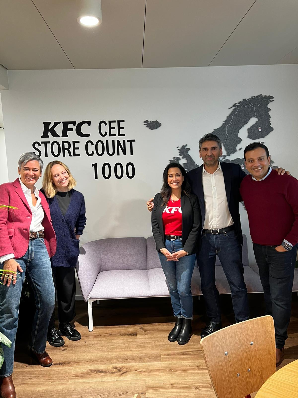 Five smiling people standing in a room with a couch and "KFC CEE store count 1000" on the wall behind them.