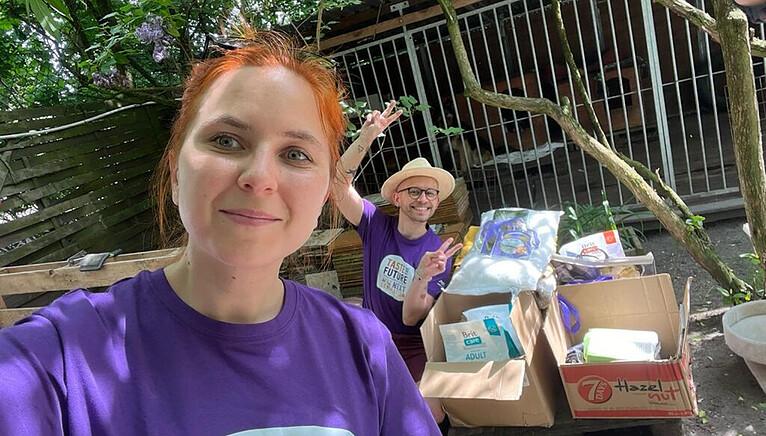 Karol in the background of a selfie with another teammate. Packed boxes of supplies on the ground next to them.