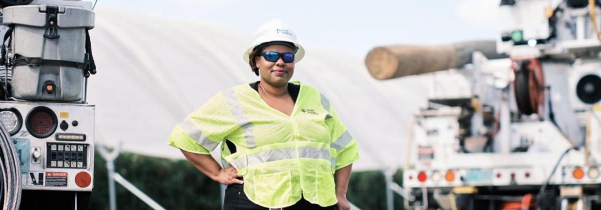 LaQuitta Ghent next to large equipment, wearing high-vis vest at hard hat. One hand on a hip.