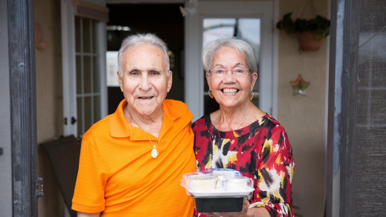 two elderly people on a front porch, one holding a packaged meal