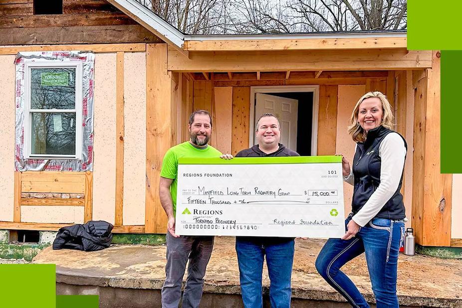 Three people holding a large check, a home being re-built behind them.