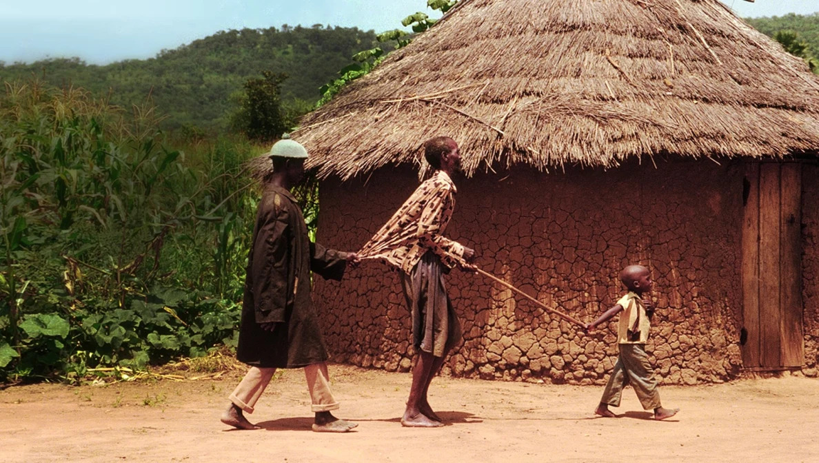 a child leads a person followed by another. a grass hut behind them