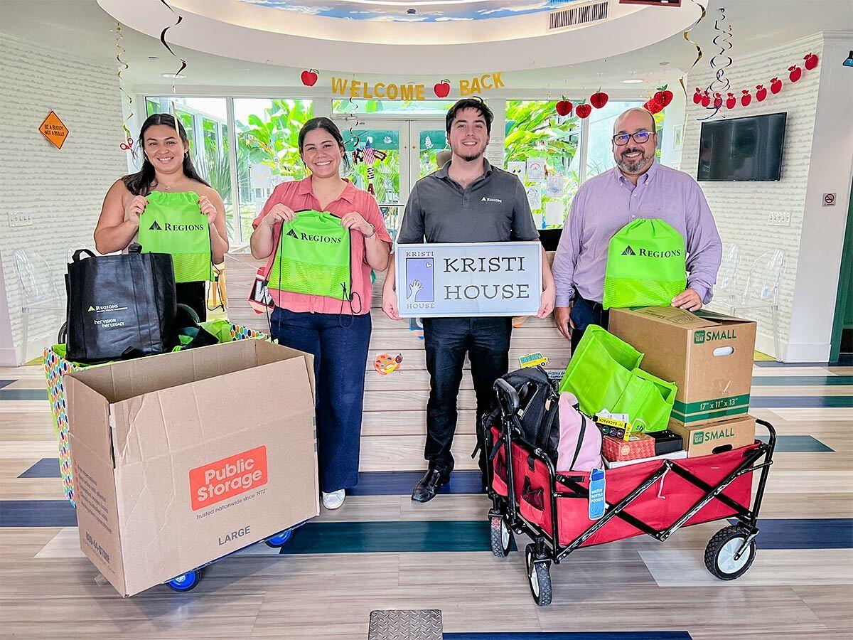 Four people posed with Regions bags, and a sign for Kristi House. Boxes of supplies in front.